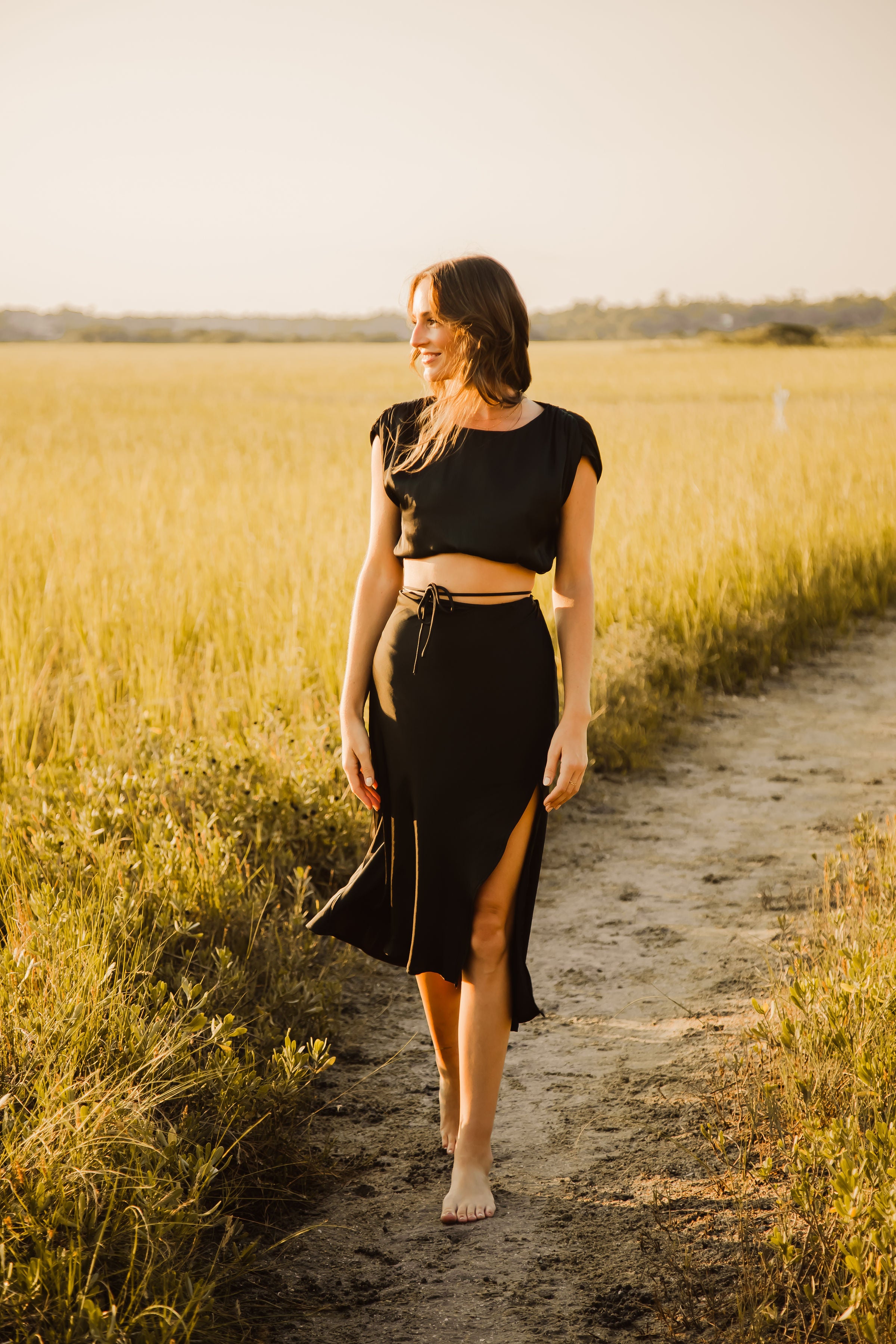 A woman walking on a dirt path in a field, dressed in a black outfit with bare feet.